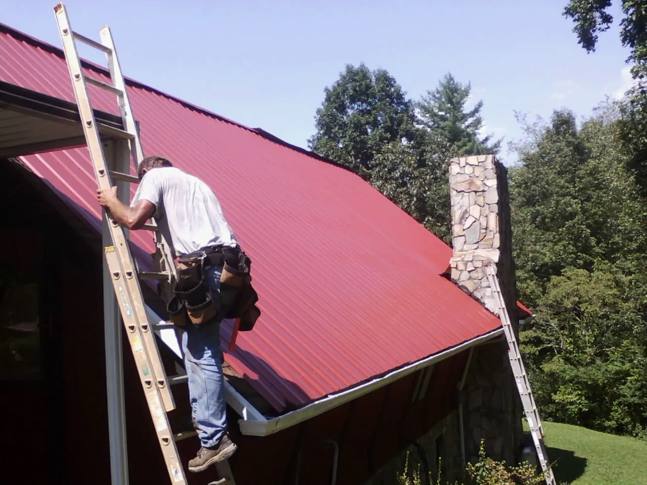 Roofer on a ladder beside a red metal roof and stone chimney — Soffit & Fascia Repair by Roofing Brothers in Warrenton